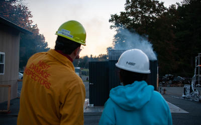Two NIST researchers, wearing hardhats and protective jackets, watch a small shed burn as part of a setudy.